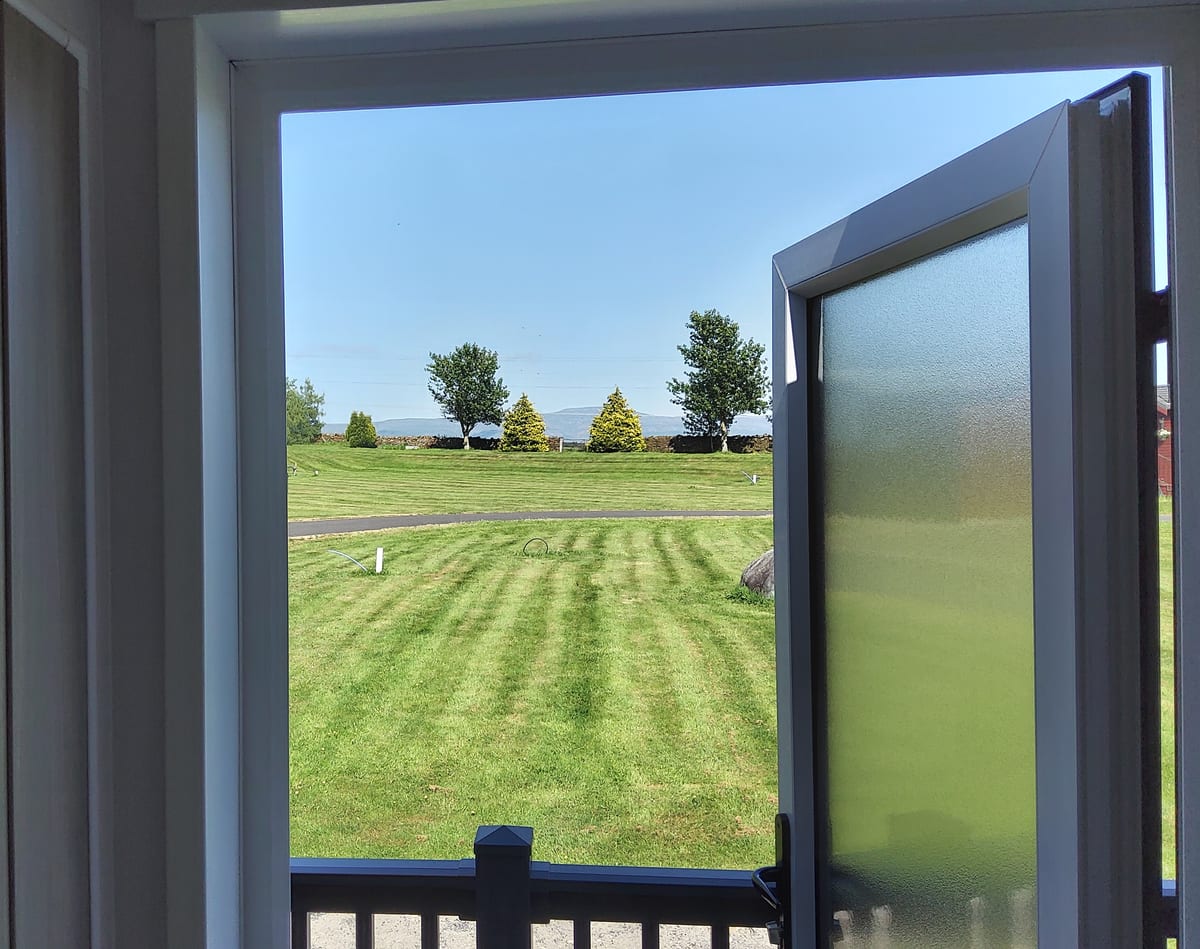 The view out of the door of a lodge at Foxfield Park: mown grass, a hedge line, trees and the fells on the horizon.